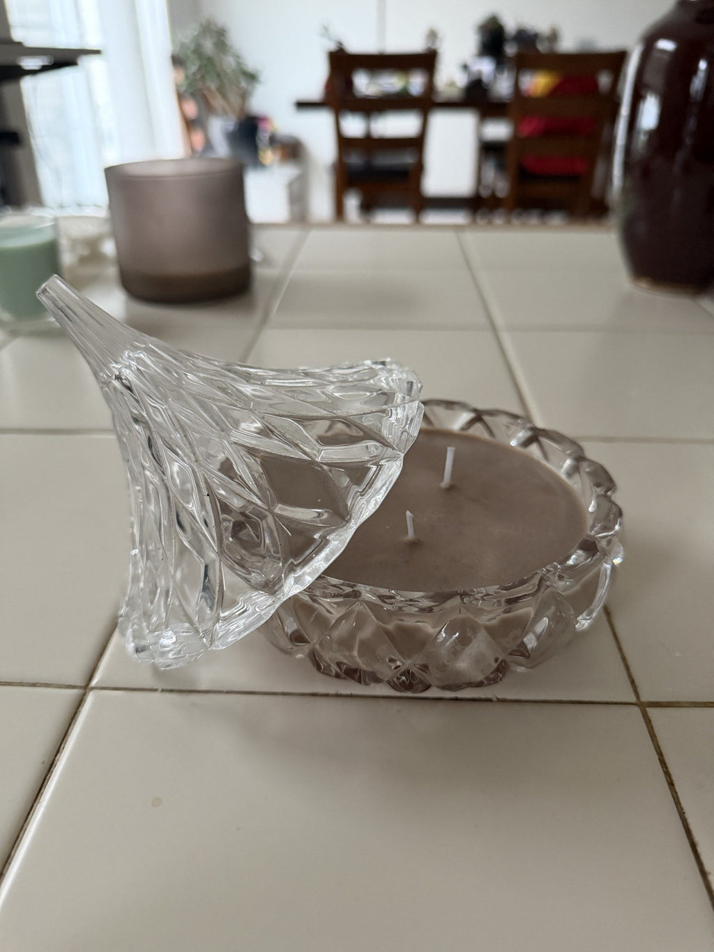 Decorative clear glass dish filled with brown candle wax, with its pointed glass lid resting beside it.